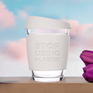 A reusable coffee cup with white lid and sleeve, standing on a beige wood block, with many tulips around it, with a background of light blue sky and pink clouds, top sunlight, front view.