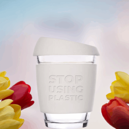 A reusable coffee cup with white lid and sleeve, with tulips around it, with a background of light blue sky and pink clouds, top sunlight, front view.